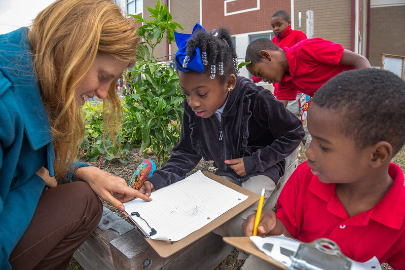 A teacher with a notepad engages with students in learning garden