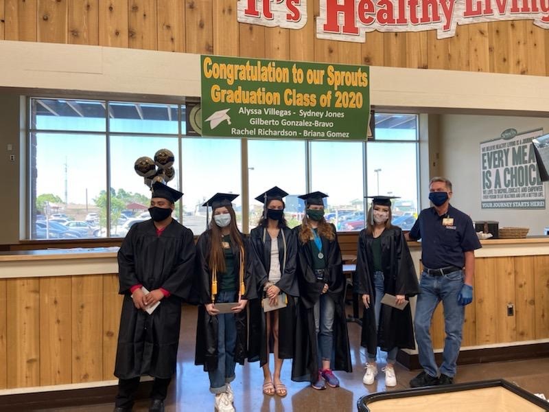 five team members at the front of a Sprouts store in caps and gowns aside the store manager and a sign congratulating them