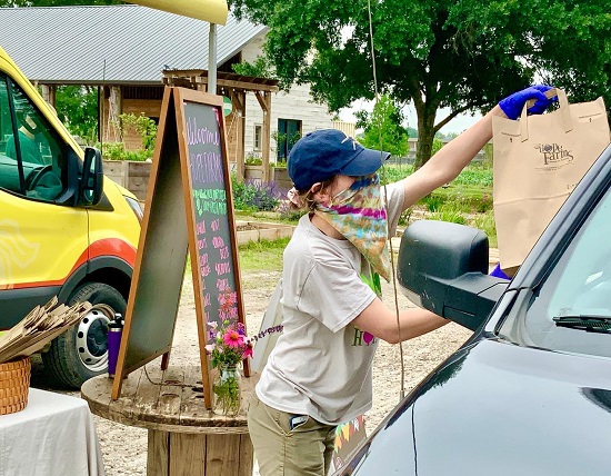 A woman at Hope Farms hands a brown paper bag of produce to a farmer's market customer, through their car window.