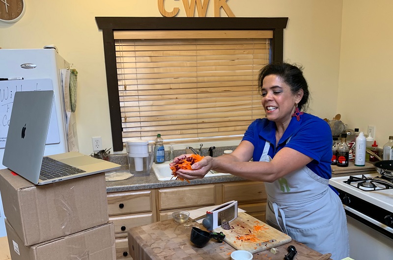 An online cooking instructor holds up carrots for students watching the class.